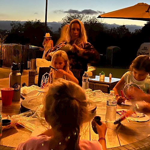 A woman and several children are gathered around a table outside in the evening, engaged in an activity, with drinks and snacks on the table.