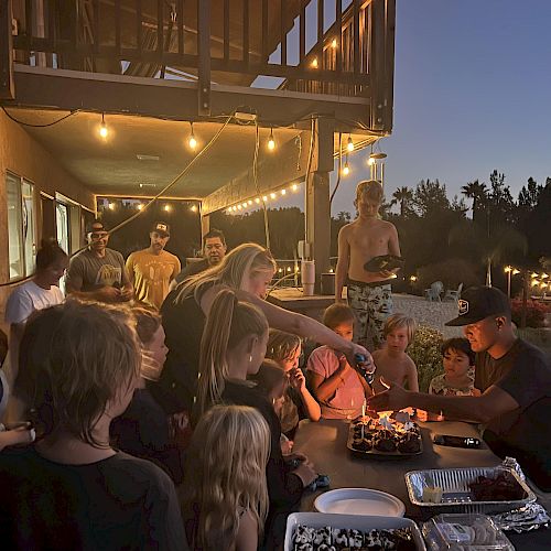A group of people gathers outside under string lights, celebrating with food and a cake. The setting is a deck with a warm, ambient evening atmosphere.