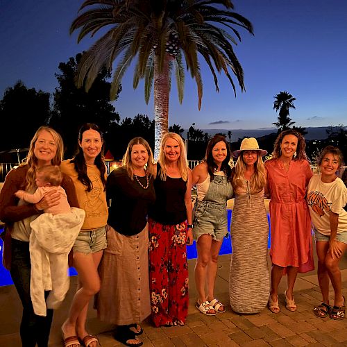A group of women stands together outdoors at dusk with a pool and palm trees in the background, all smiling for a photo.