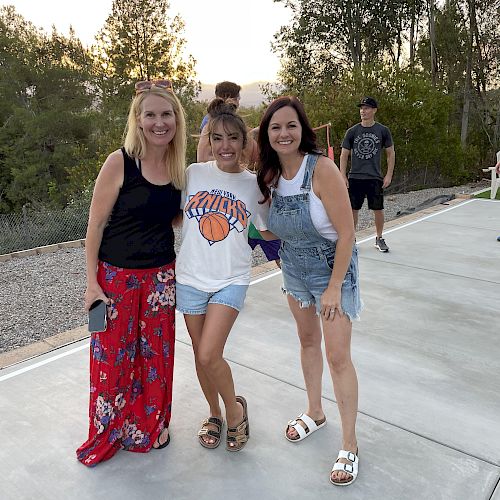 Three women are posing and smiling outdoors on a paved path with trees in the background, while a few people are walking behind them.