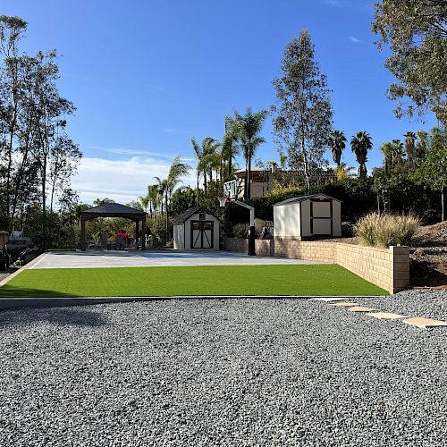 The image shows a landscaped area with a gravel driveway, green artificial turf, two sheds, stone pathway, and surrounding trees and plants under a blue sky.