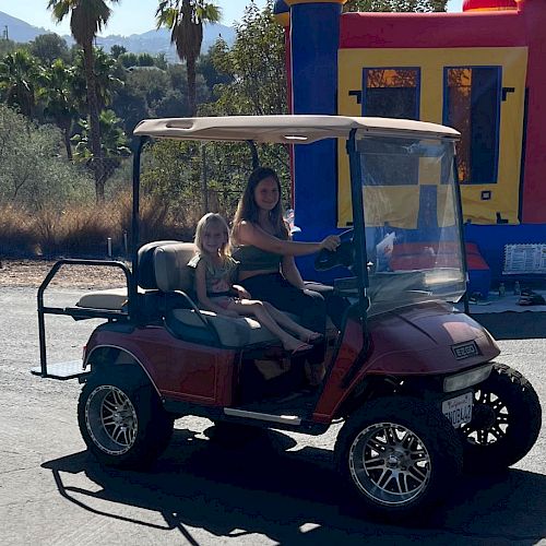 A golf cart with two people, parked in front of a colorful bounce house, with trees and mountains in the background on a sunny day.