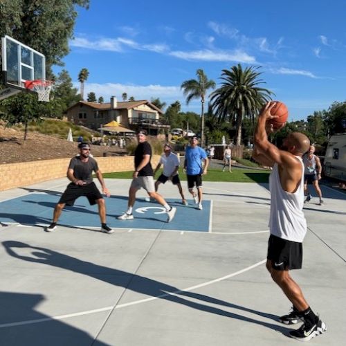 A group of people playing basketball outdoors on a sunny day, with one person preparing to take a shot while others play defense on the court.