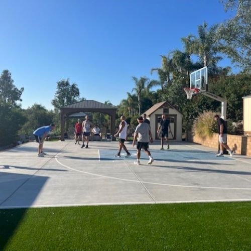 A group of people are playing basketball on an outdoor court surrounded by greenery and a clear sky.