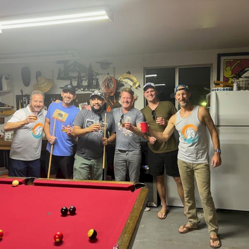 A group of six men standing around a red pool table, holding drinks and pool cues, in a well-lit garage or game room.