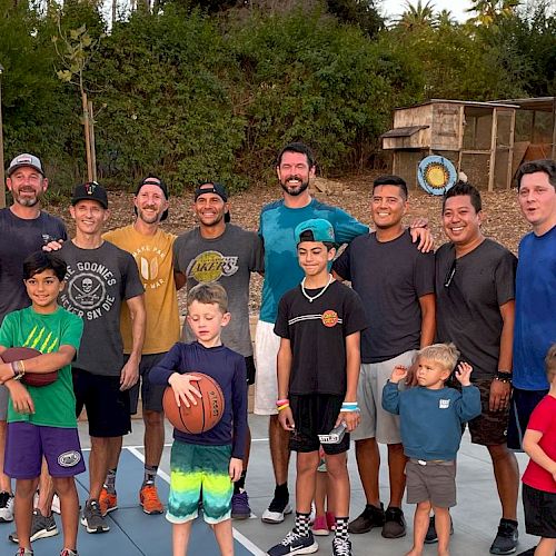 A group of people, including adults and children, are posing on a backyard basketball court while holding basketballs, smiling for the photo.