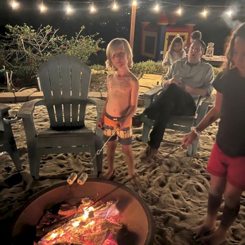 Children and adults enjoying a nighttime bonfire, with one child roasting marshmallows while others relax in chairs under string lights.