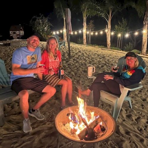 Three people sit around a campfire on sandy terrain, holding drinks. String lights and palm trees are in the background, creating a cozy ambiance.