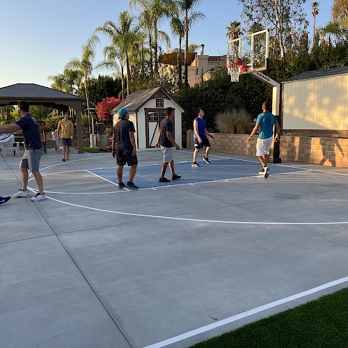 A group of people playing basketball on an outdoor court with a backdrop of trees and a shed.
