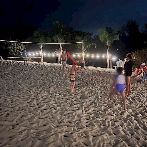 People are playing volleyball on a sand court at night, with others sitting and watching nearby under string lights and palm trees.