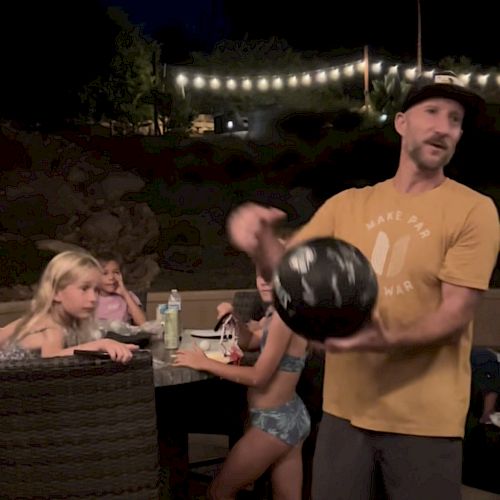 A man in a yellow shirt and cap holds a ball while beside a table with children, possibly in an outdoor setting at night with string lights in the background.