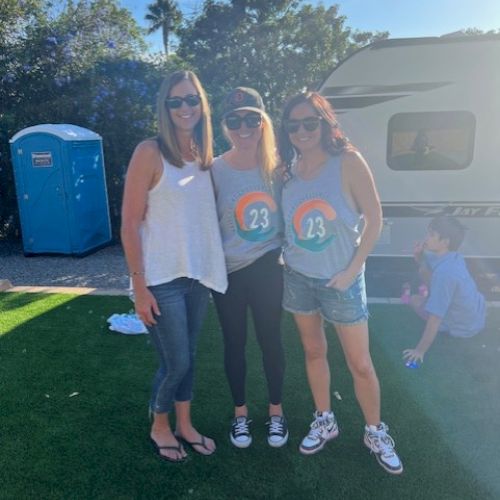 Three women stand on grass in front of an RV, two wearing matching shirts. A portable toilet is nearby, and a child plays in the background.