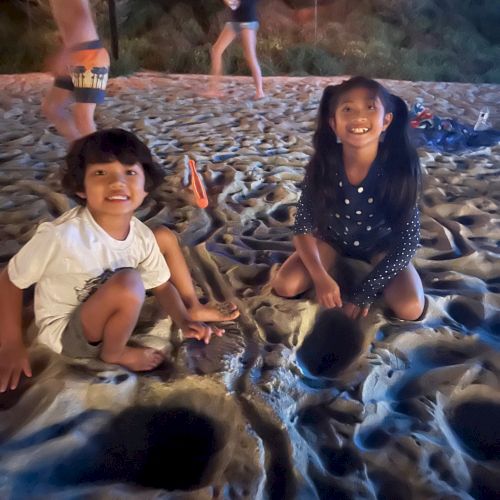 Two children are sitting on the sand and smiling at the camera at night. Other people and lights are visible in the background.