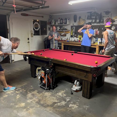 Four people are in a garage playing pool on a table with a red felt surface. Various tools and items are hung on the walls.
