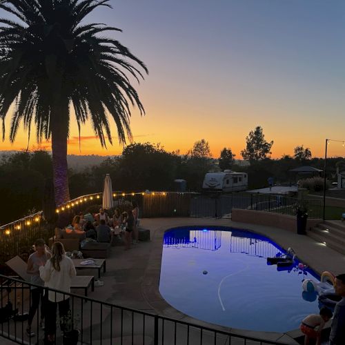 People are gathered around a lit pool at sunset with a palm tree, lights, and a trailer in the background.