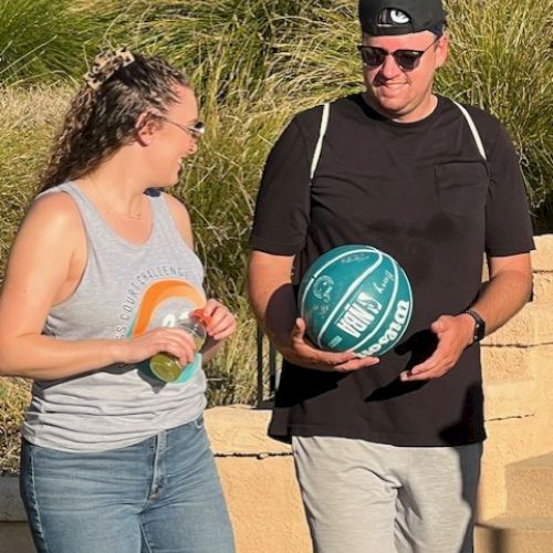 Two people walking outdoors, one holding a green basketball. Both are casually dressed and appear to be engaging in conversation.